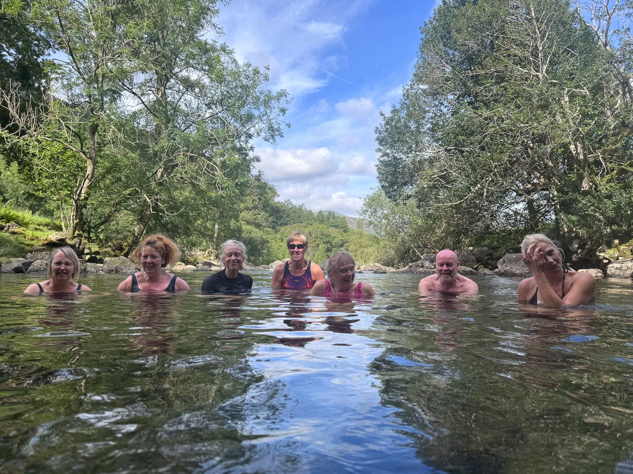 Seven people are standing in a river surrounded by trees and nature, smiling and enjoying a sunny day.