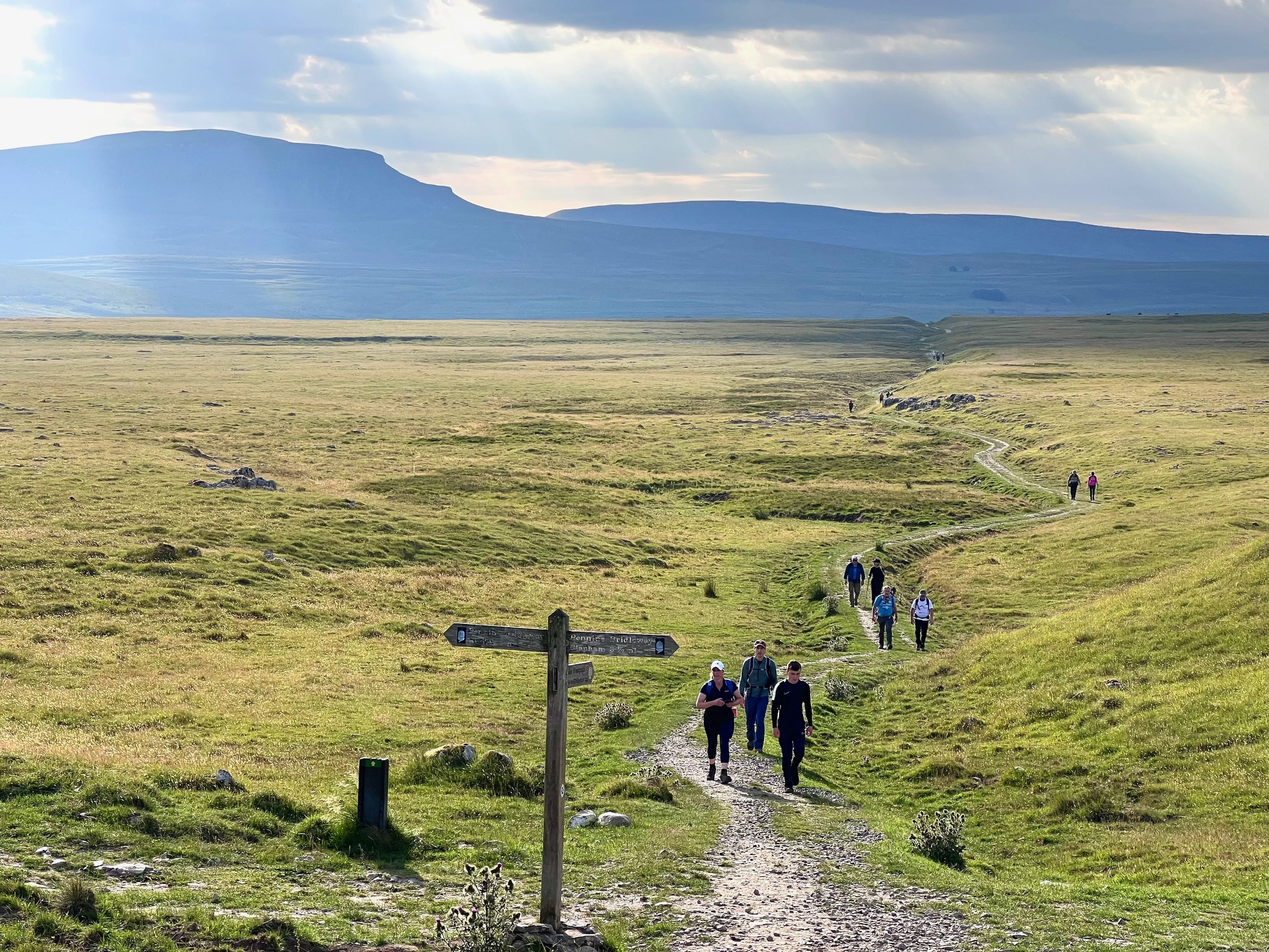 Hikers on a grassy trail in a hilly landscape under a cloudy sky with sunbeams.