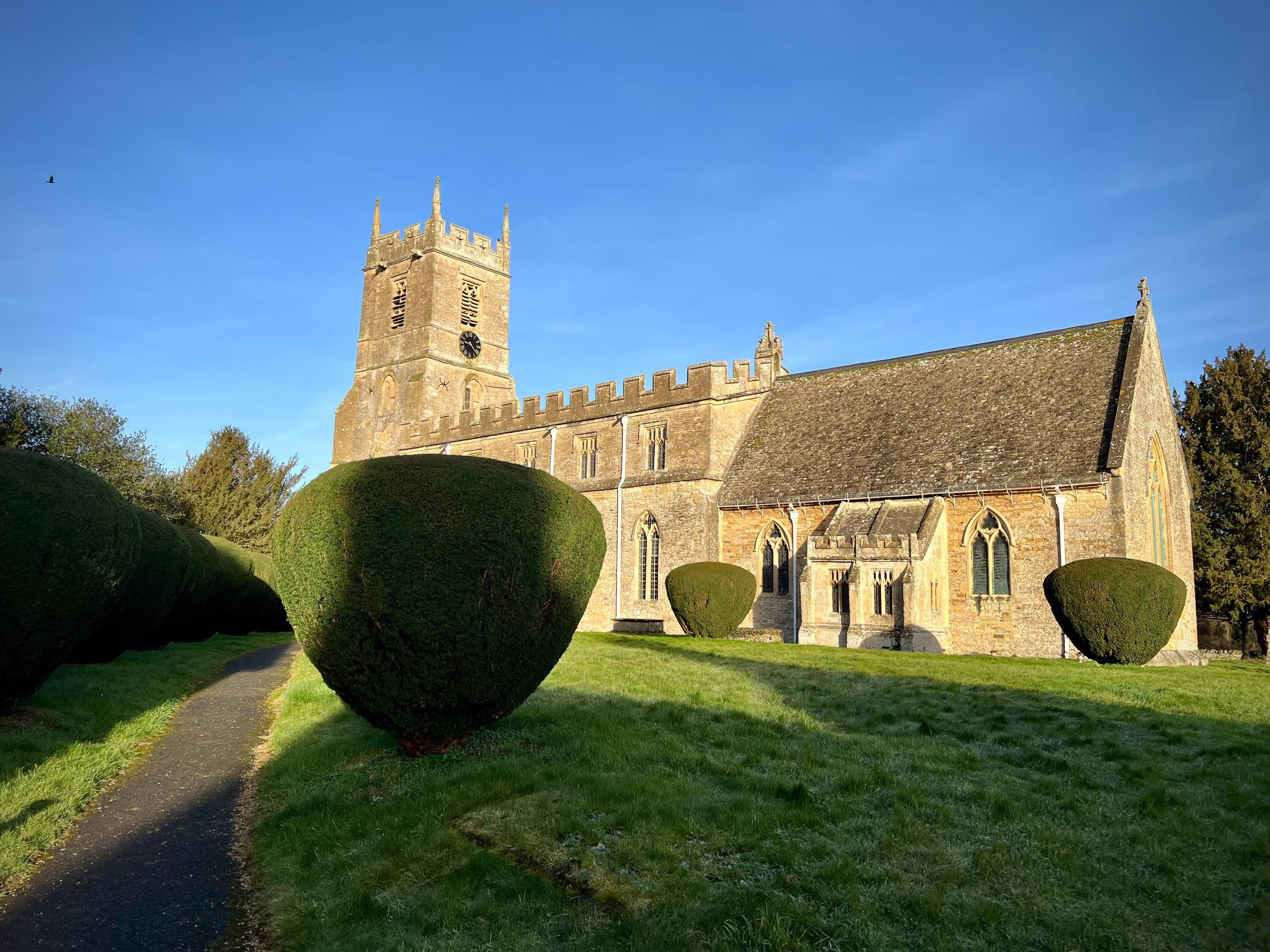 Medieval stone church with bell tower, arched windows, and manicured hedges, under a clear blue sky.
