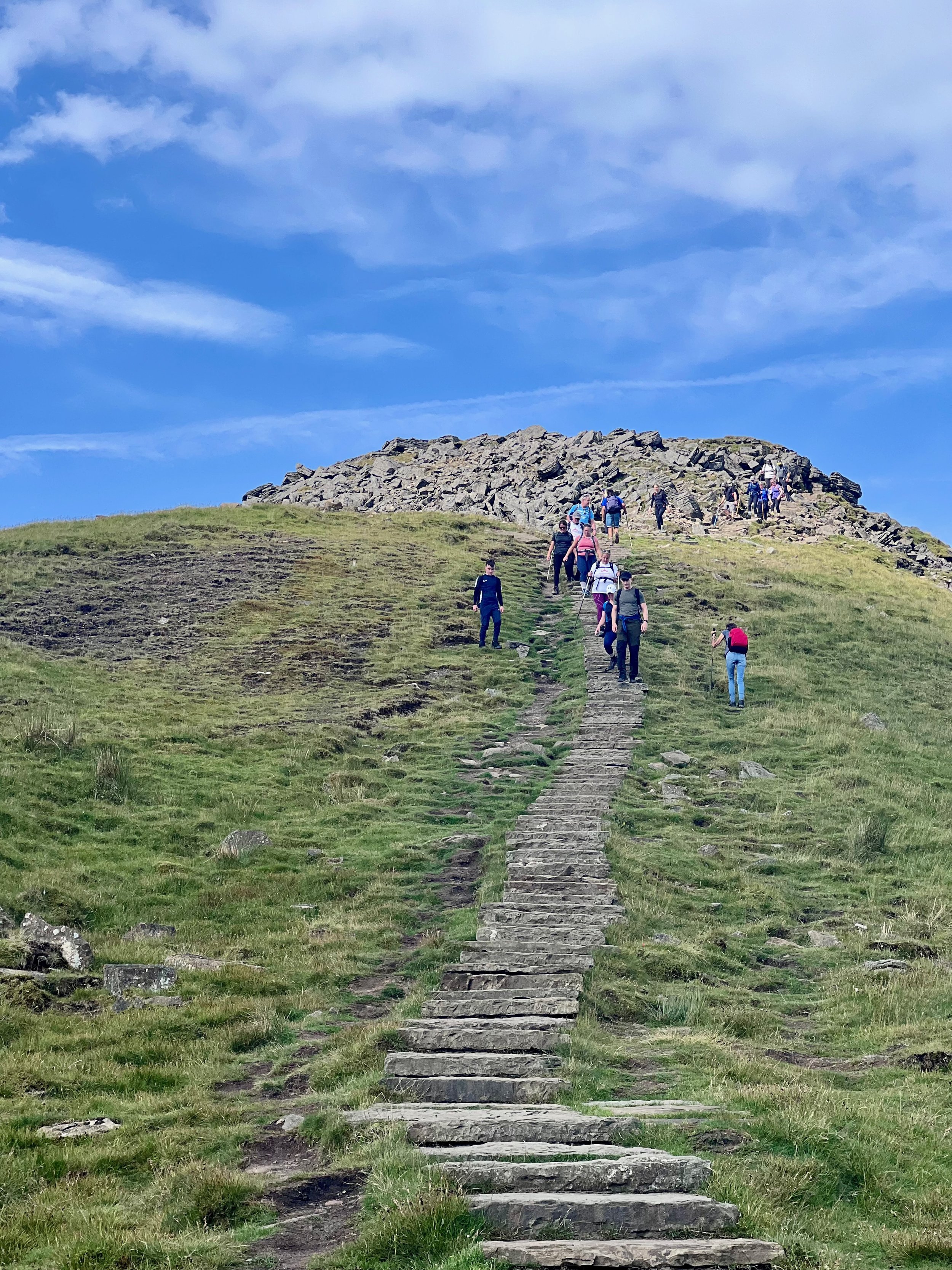Hiking trail with stone steps leading up a grassy hill, people walking, blue sky with clouds
