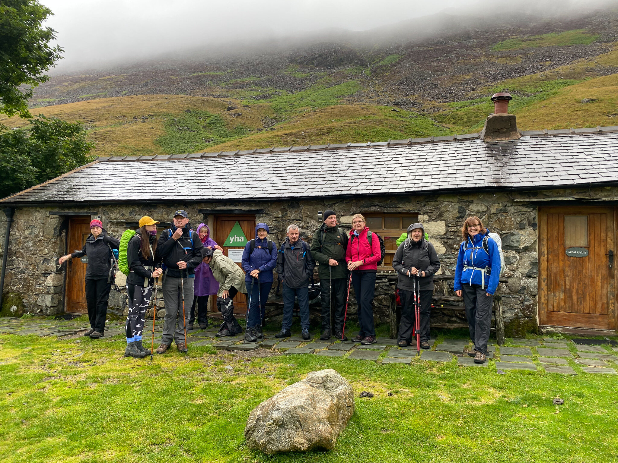 Group of hikers in outdoor gear standing in front of a stone building with a "YHA" sign, set against a misty hillside.