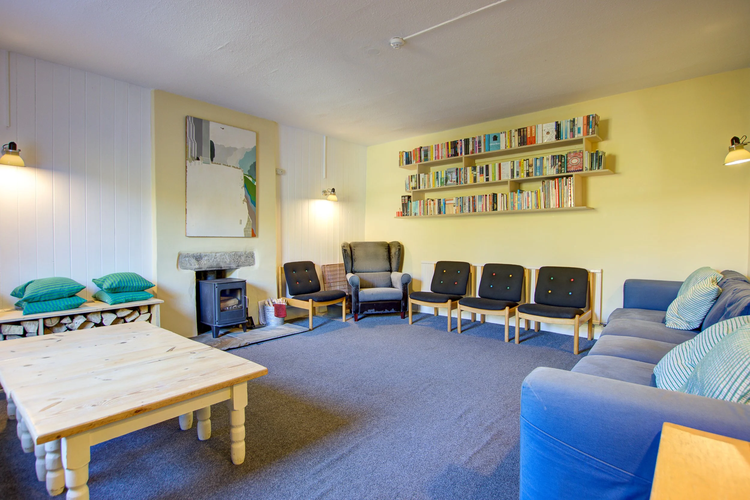 A cozy living room with a wood-burning stove, a framed artwork, a yellow wall with shelves filled with books, and various chairs and a couch arranged for seating.