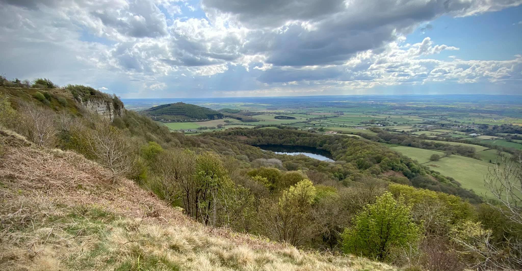 Scenic landscape view from a hillside, featuring green fields, a small lake surrounded by trees, and a cloudy sky.
