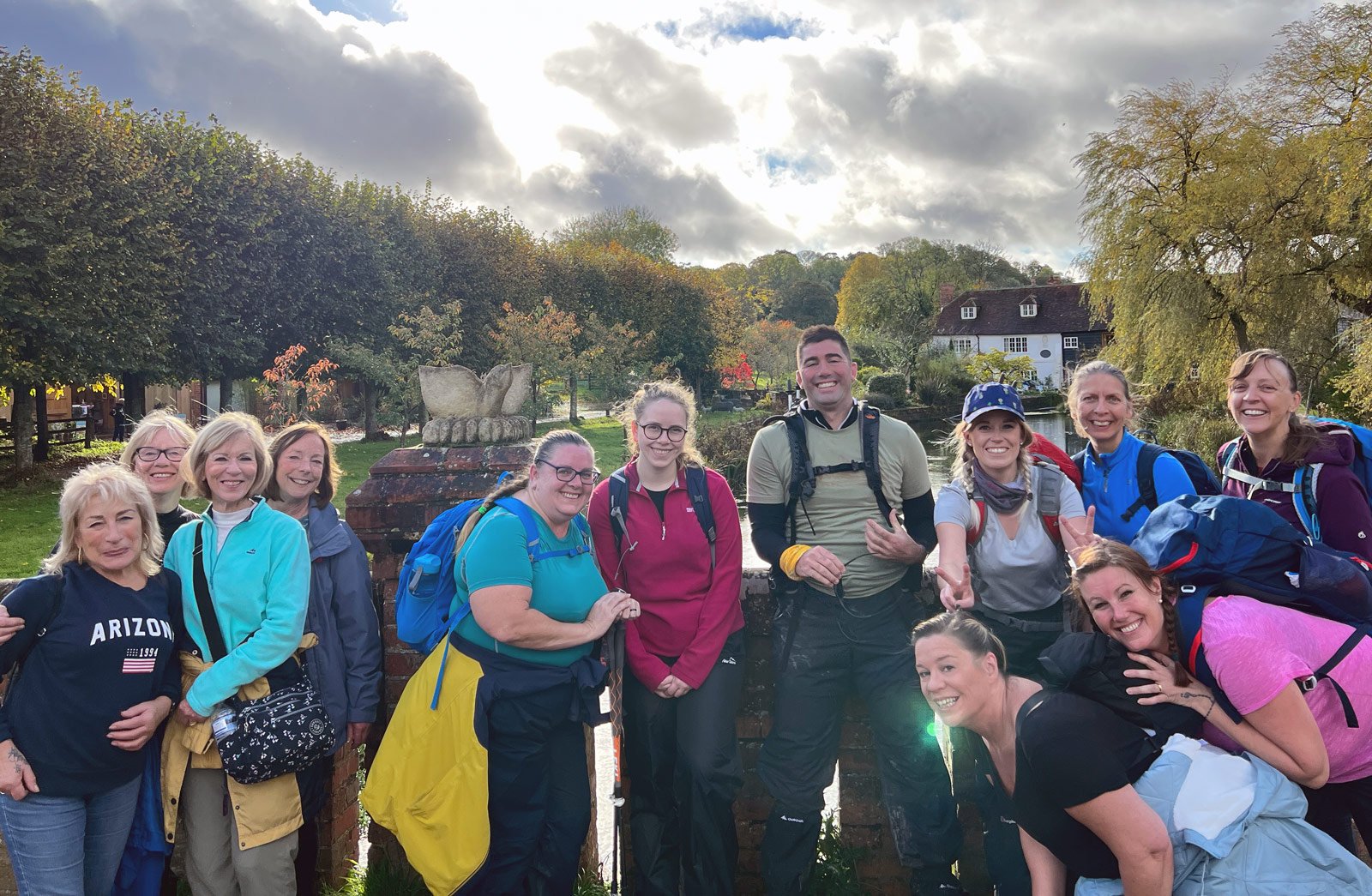 Group of people with backpacks smiling outdoors near trees and a house.