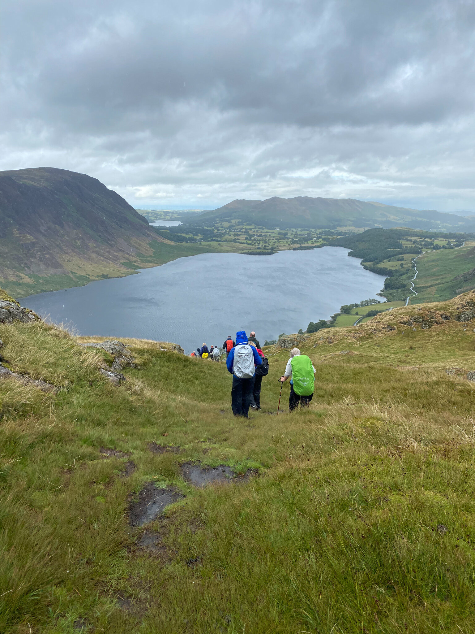 Hikers descending a grassy hill towards a large lake, surrounded by rolling green hills and cloudy skies.