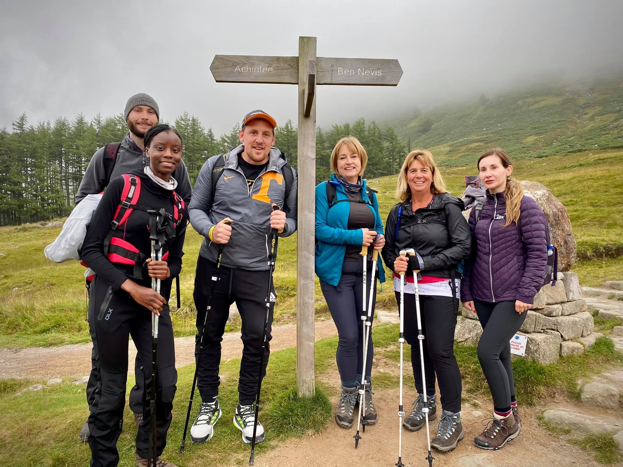 A group of six hikers are posing in front of a sign with "Achintee" and "Ben Nevis" directions. They are standing on a path surrounded by grass and trees. The hikers are equipped with backpacks and trekking poles, wearing outdoor gear suitable for hi