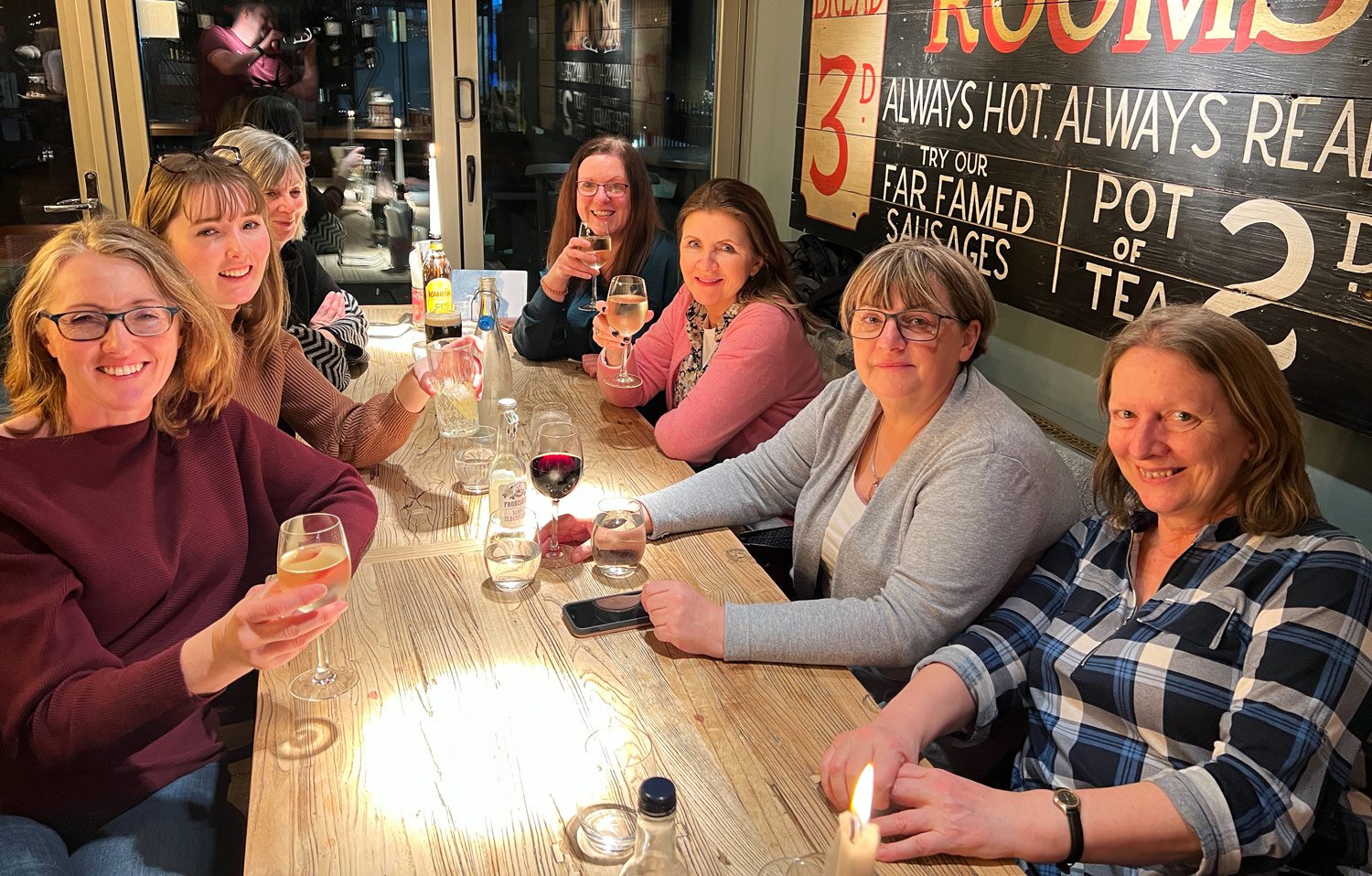 A group of women sitting at a wooden table in a restaurant, each holding a glass of wine, smiling for the camera. A vintage sign is visible on the wall behind them.