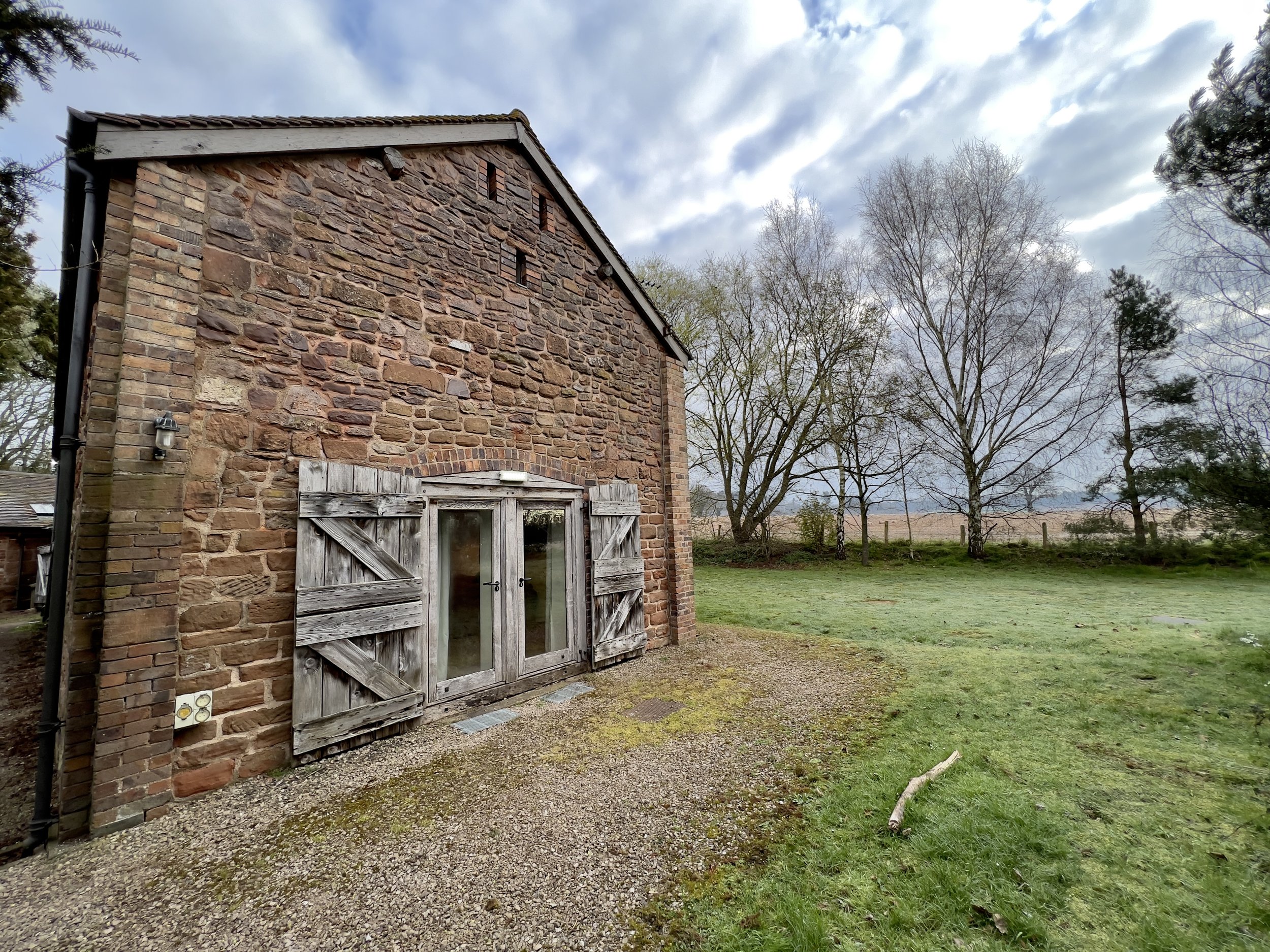 Old stone building with wooden shutters and a door, surrounded by grass and trees, under a cloudy sky.