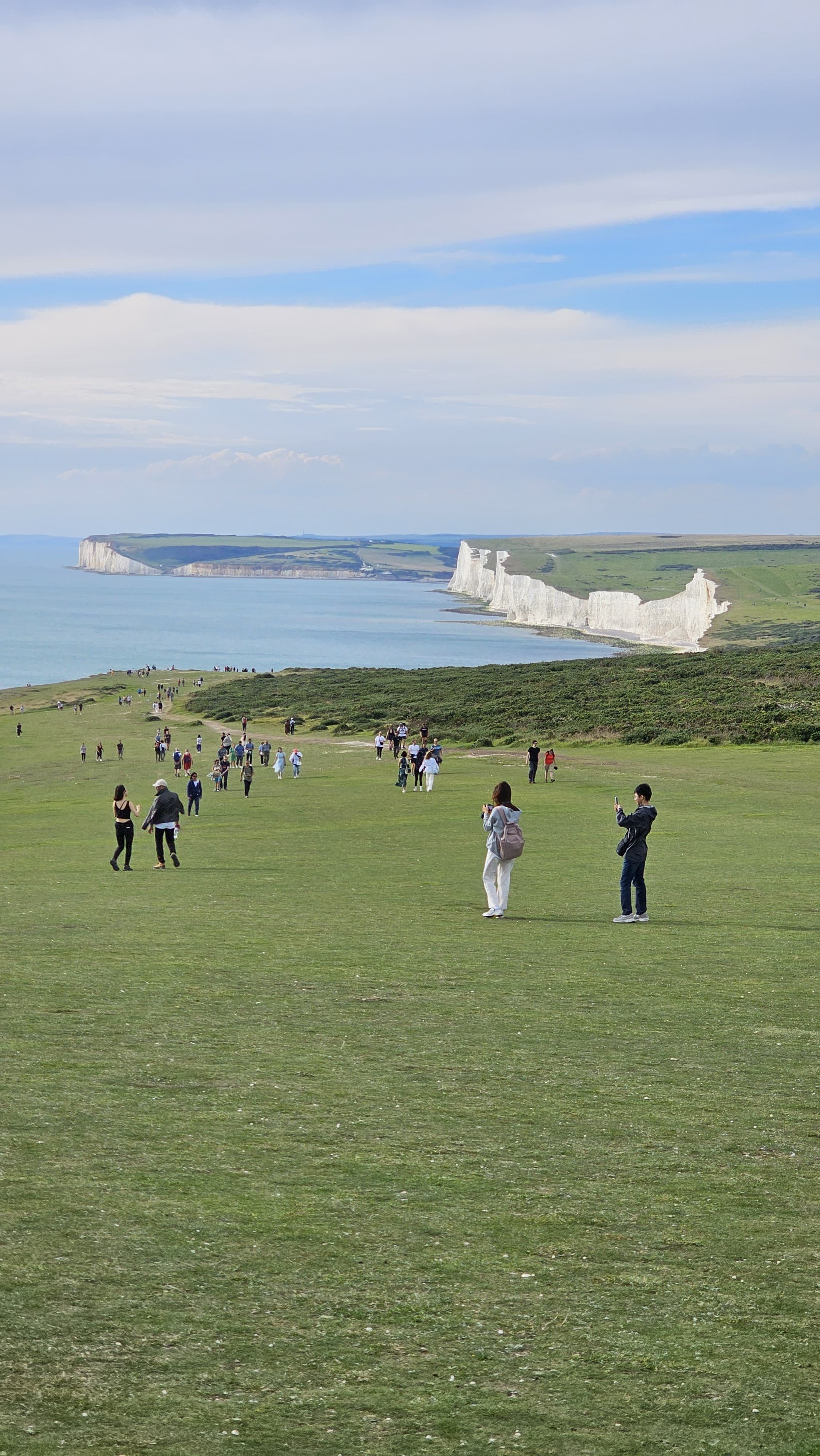 People walking and taking photos on a grassy hill overlooking the white chalk cliffs of Seven Sisters, with the ocean in the background.