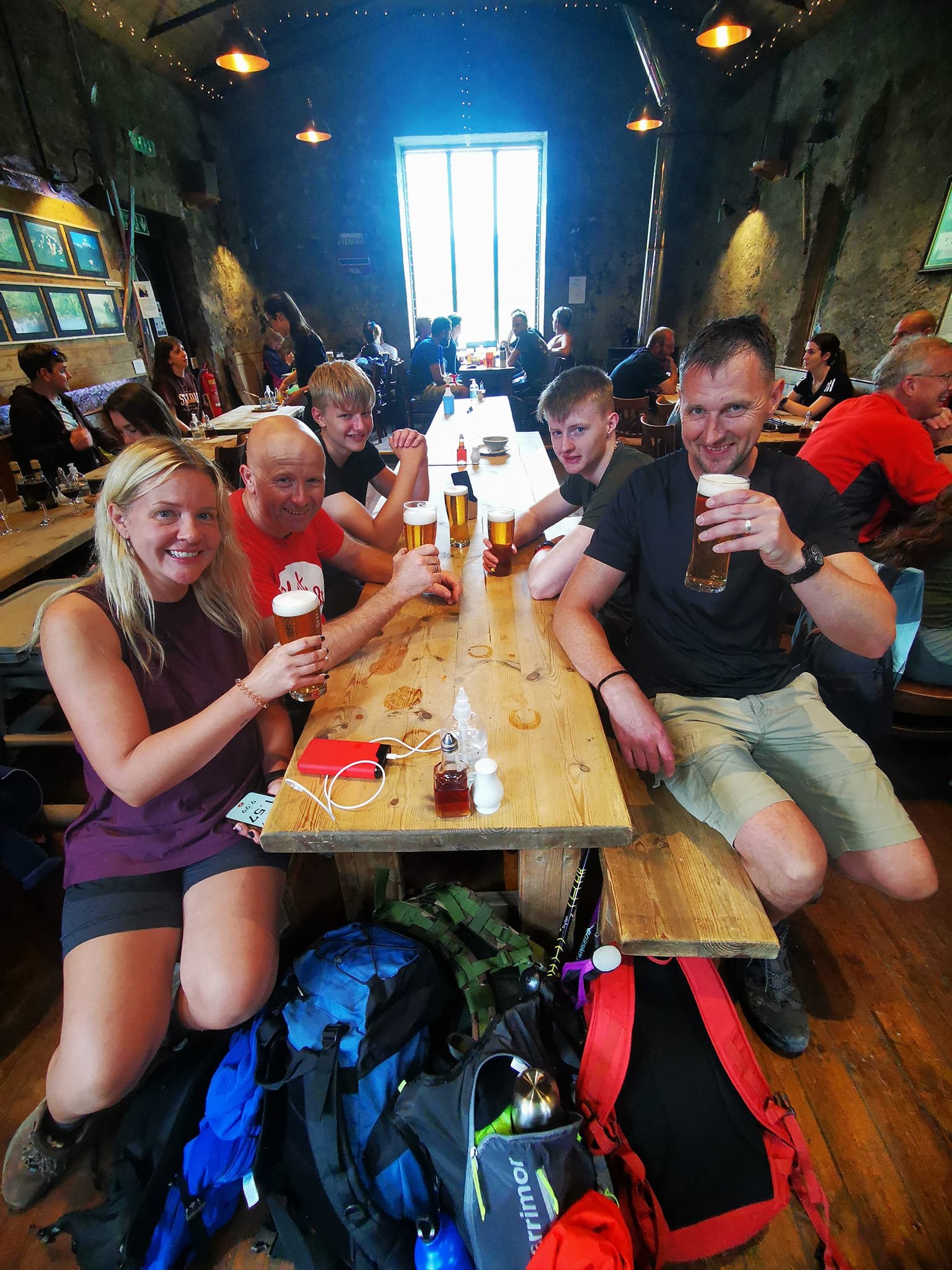 Group of people sitting at a wooden table in a pub, holding glasses of beer, with backpacks on the floor. The interior has exposed stone walls and overhead lights.