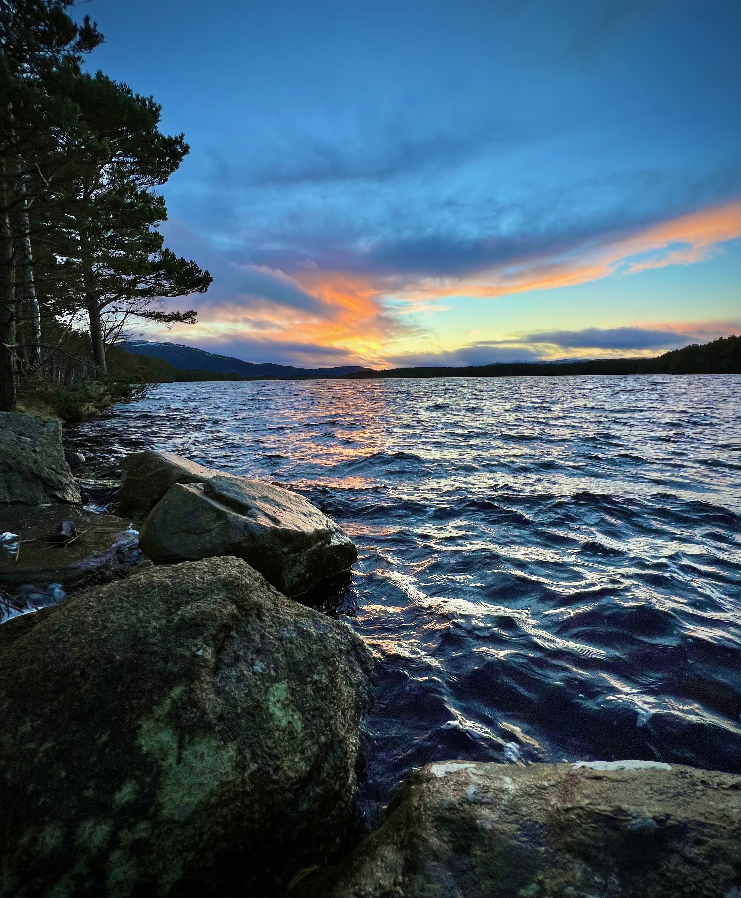 Scenic view of a lake at sunset with rippling water, distant mountains, and trees on the shoreline.