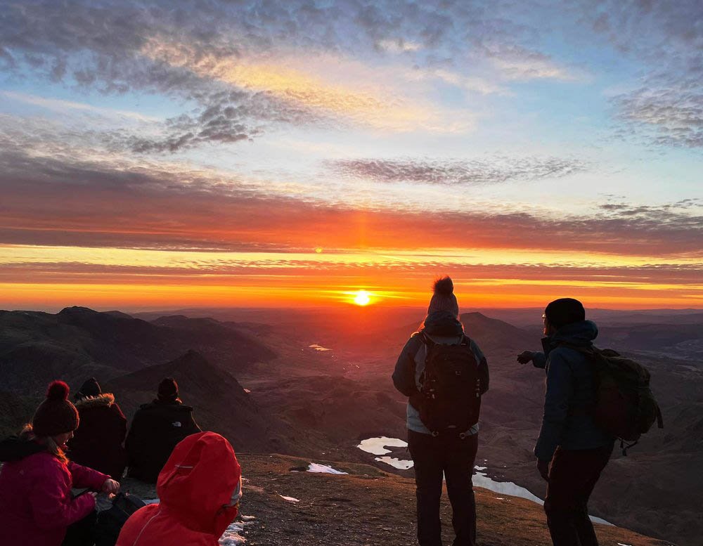 People watching a colorful sunrise from a mountaintop, with dramatic sky and distant landscape.