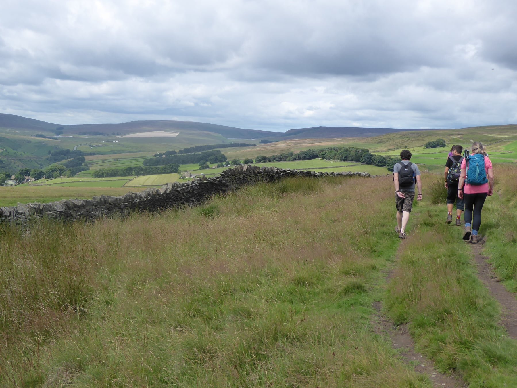 Hikers walking on a grassy trail in a rural landscape with rolling hills and a stone wall, under a cloudy sky.