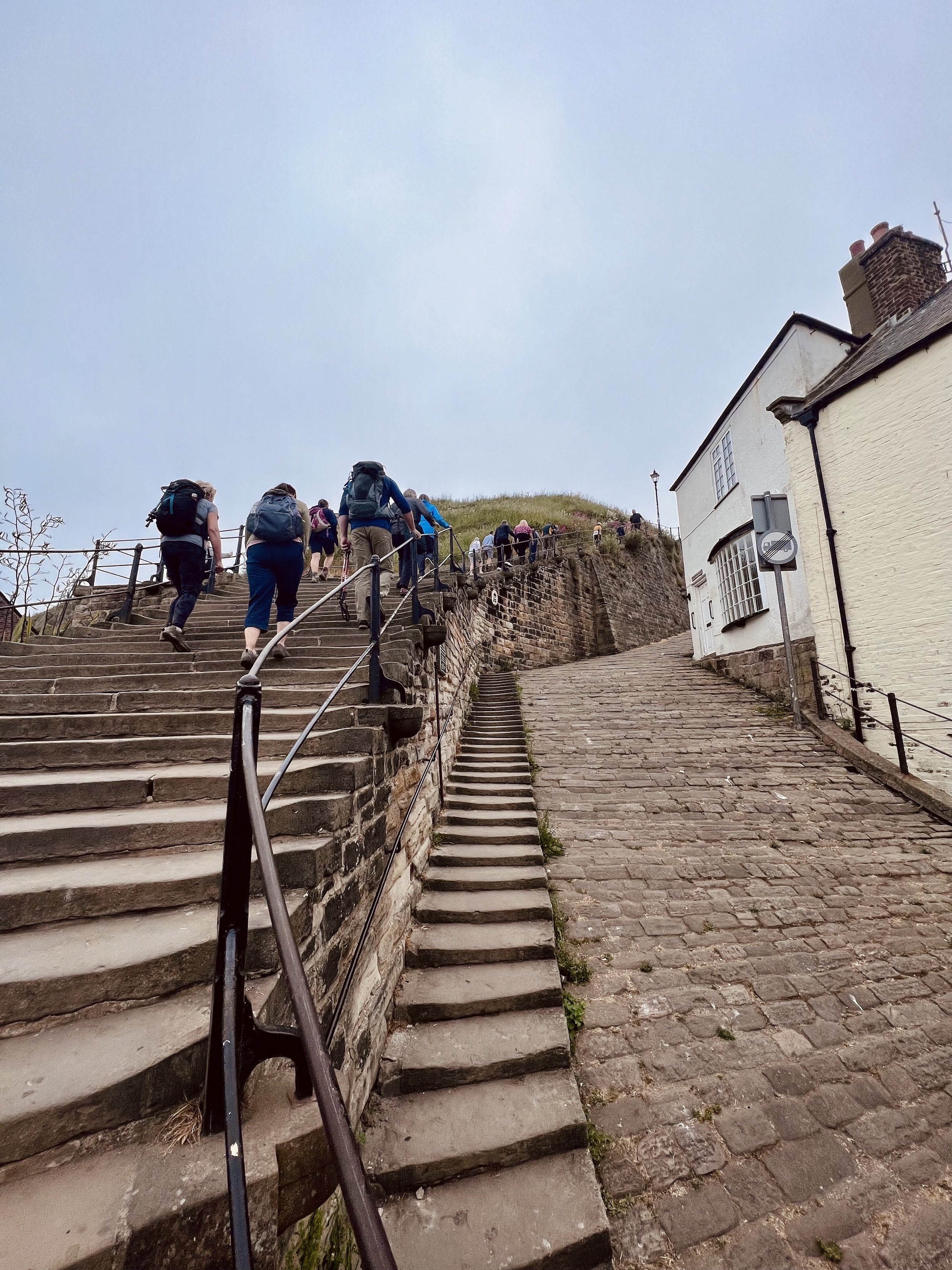People climbing an outdoor stone staircase with a wall and buildings nearby on a cloudy day.