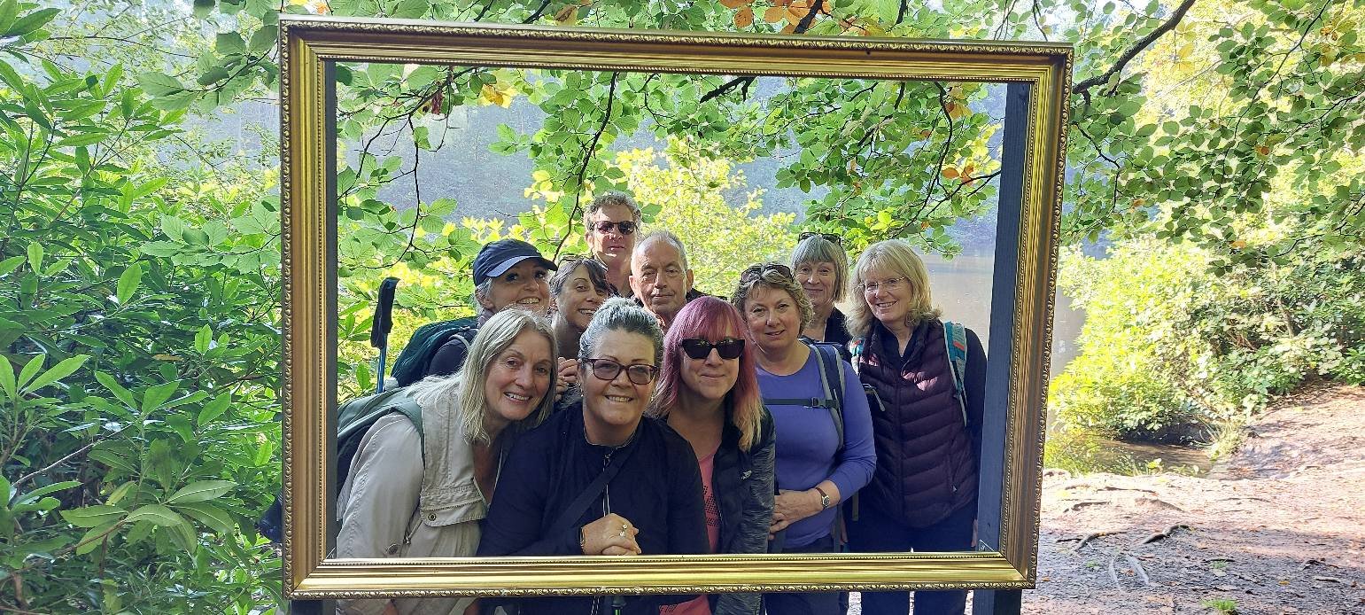 Group of people smiling in a wooded area, posing behind an empty picture frame, with lush greenery in the background.