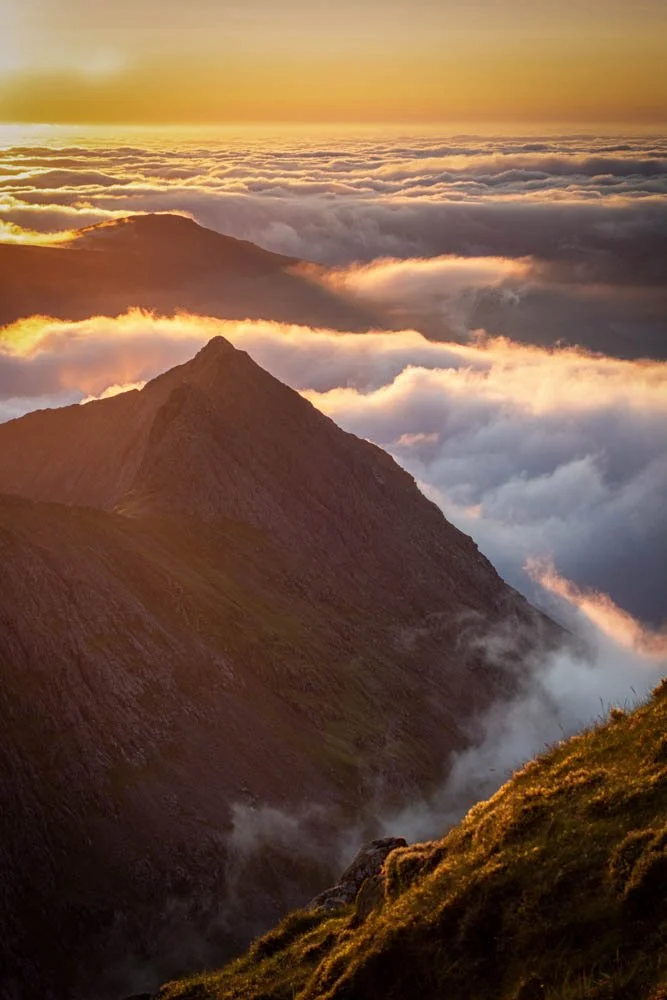 Mountain peak at sunset above clouds with warm orange glow.
