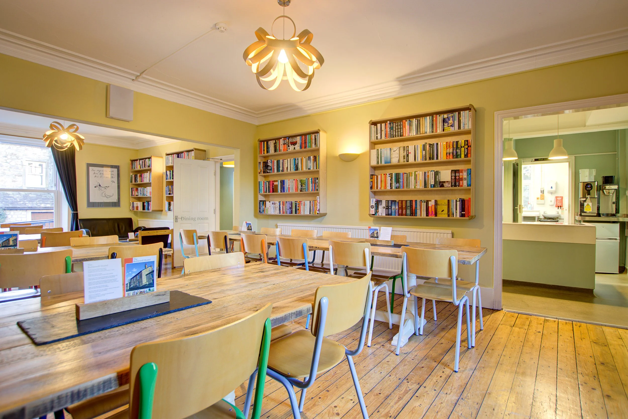 A cozy library or community space with wooden tables and chairs, bookshelves filled with colorful books on the walls, and large windows letting in natural light. Two modern light fixtures hang from the ceiling.