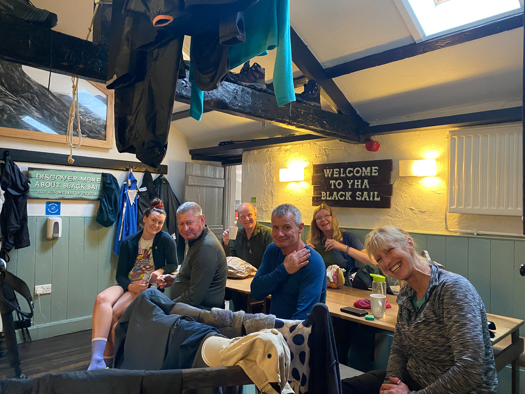 Group of smiling people sitting at a table inside a cozy room with a sign "Welcome to YHA Black Sail" on the wall, clothing hanging overhead.