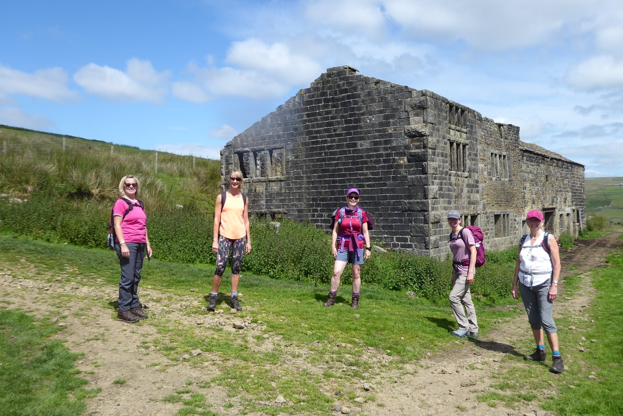 Five women hiking near a historic stone building in a rural landscape with grass and a blue sky.