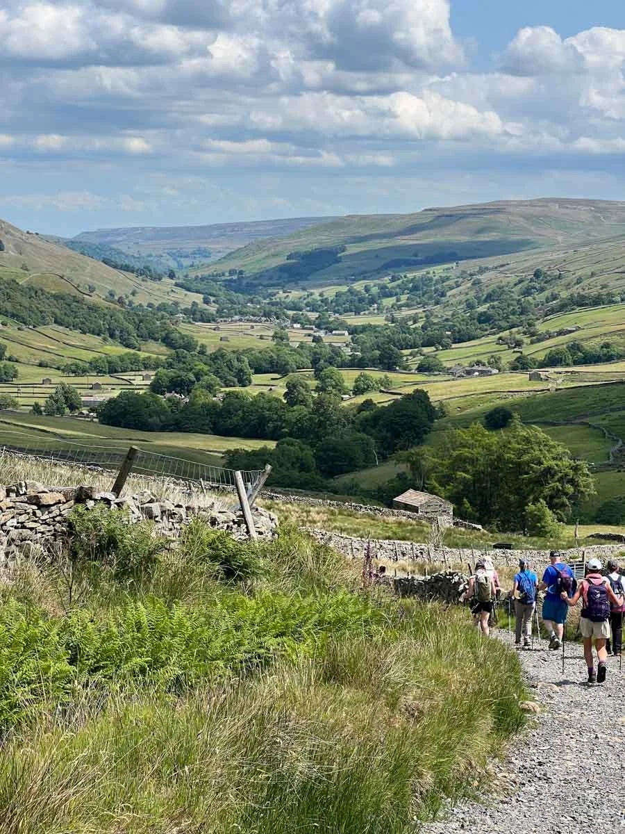 Group of hikers walking on a trail through a scenic, green valley with hills and clouds in the background.