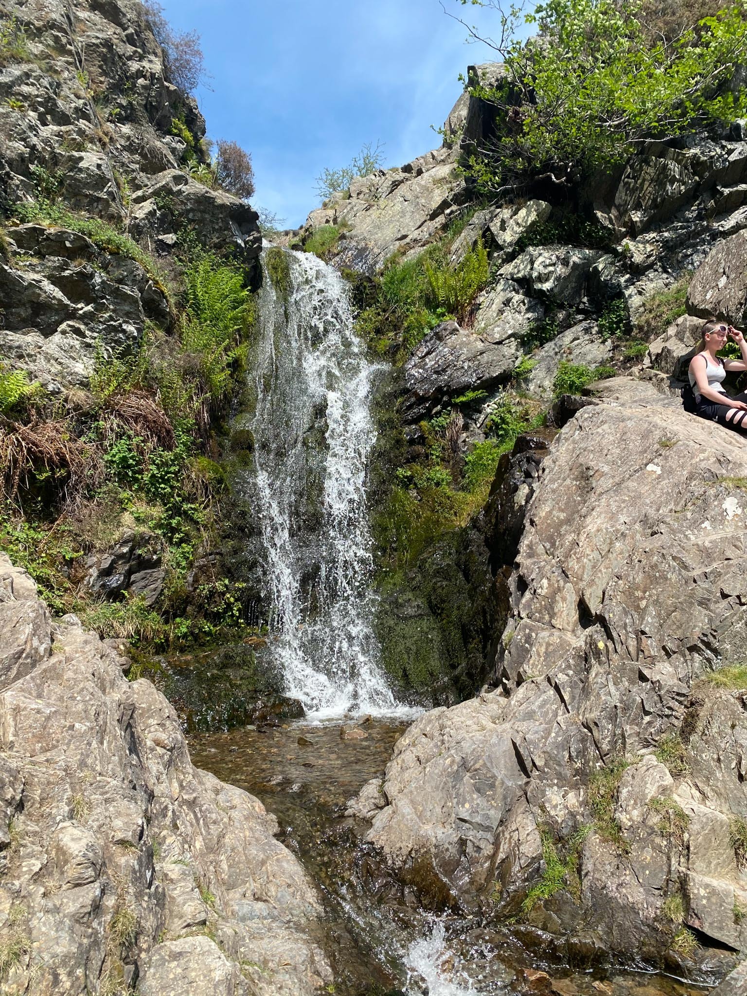 Waterfall flowing down rocky cliff surrounded by greenery and rocks, with a person sitting on a rock to the right under a sunny sky.