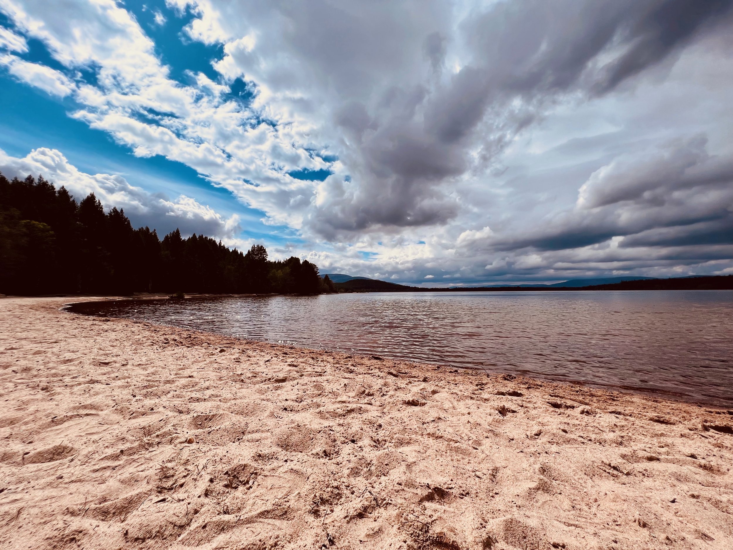 A sandy beach with a wide expanse of water under a dramatic sky filled with clouds. In the background, there are silhouettes of trees and distant hills.