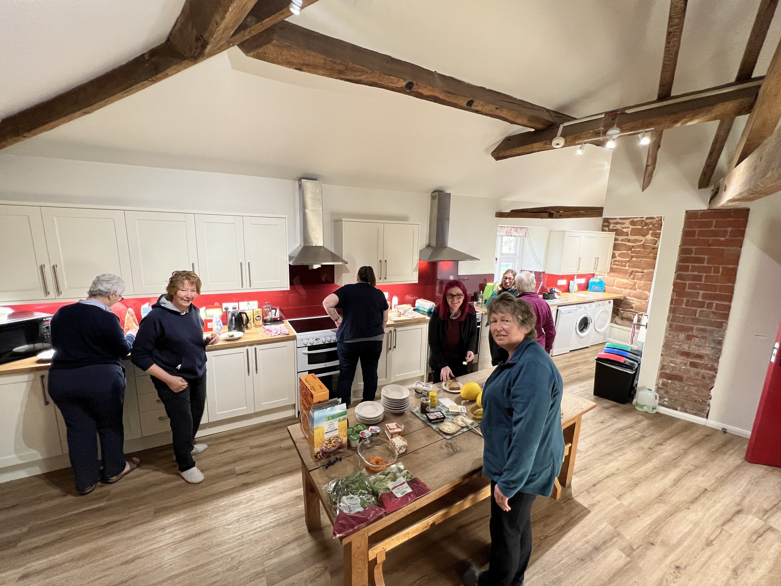 A group of people in a kitchen preparing food. The kitchen features white cabinets, red backsplash, and wooden beams on the ceiling. A wooden table in the center is filled with ingredients, snacks, and kitchenware.