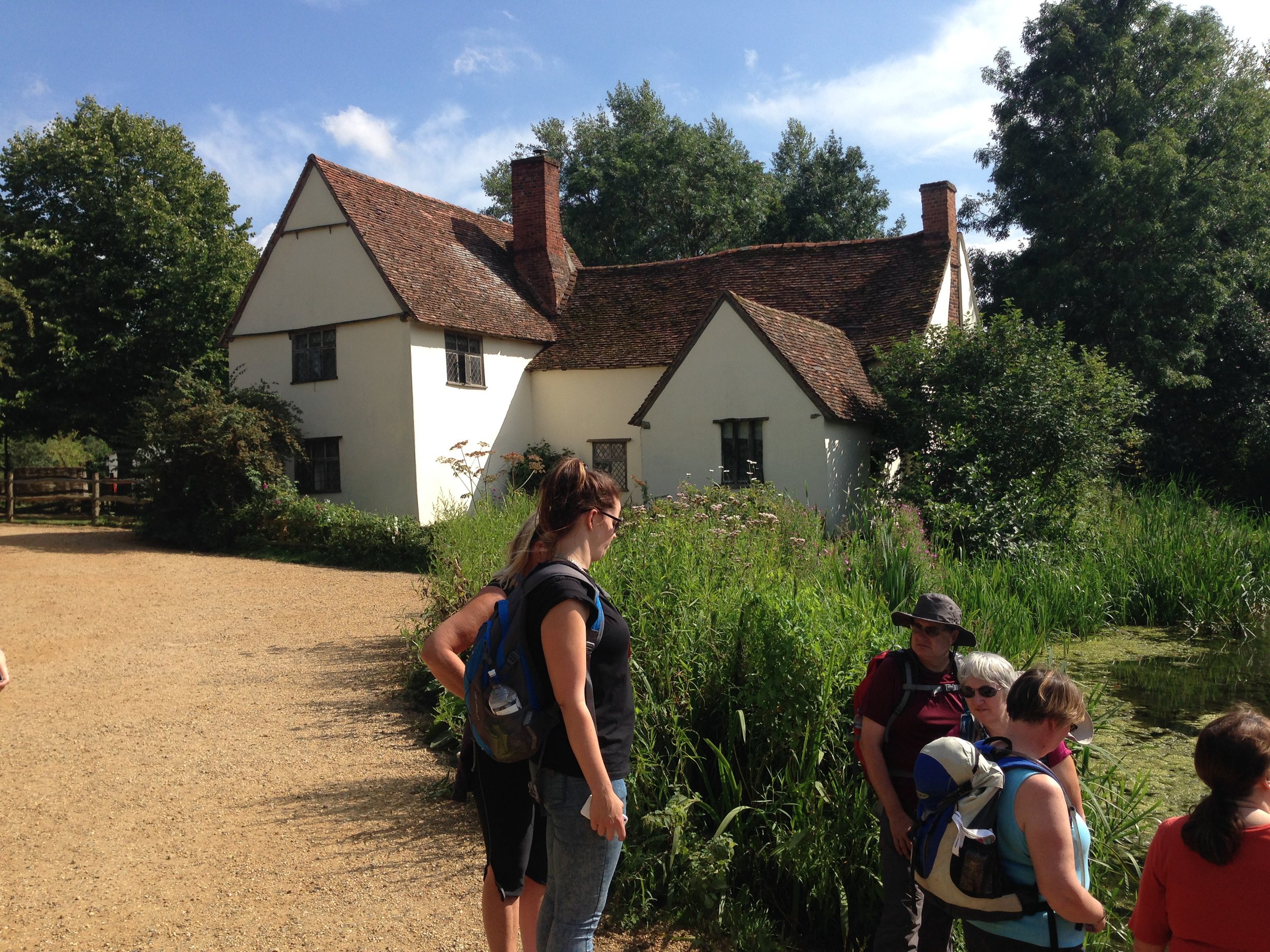 Group of hikers standing near a traditional English cottage surrounded by greenery with trees and blue sky in the background.