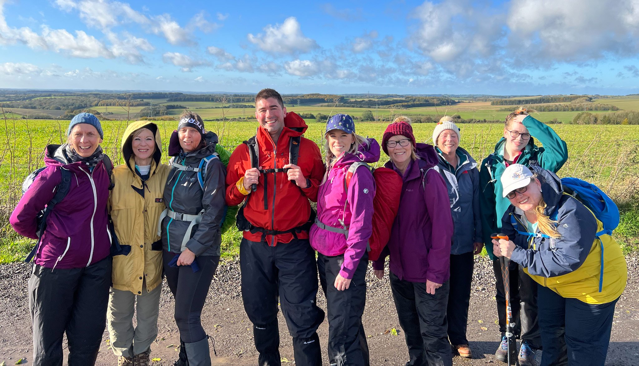 Group of people wearing colorful outdoor clothing, standing on a rural path with fields and a blue sky in the background.