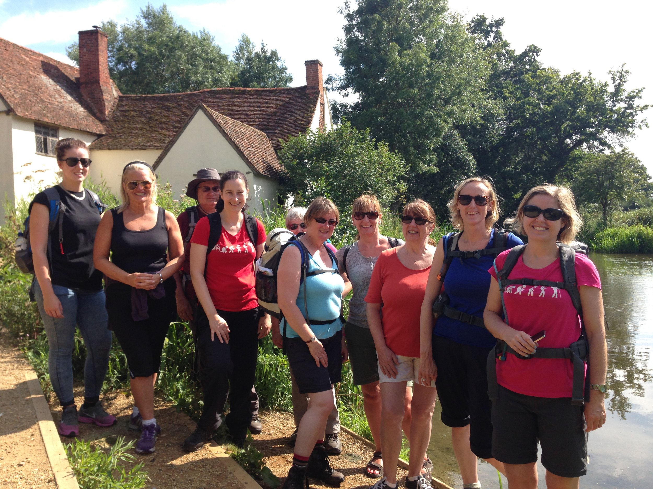 A group of women smiling outdoors by a house and a pond. They are wearing casual hiking attire and sunglasses, with trees in the background.