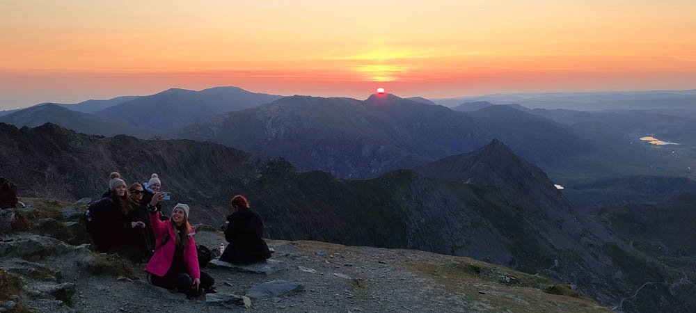 A group of people sitting on a rocky mountain peak taking a selfie during a sunset, with a vast landscape and mountains in the background.