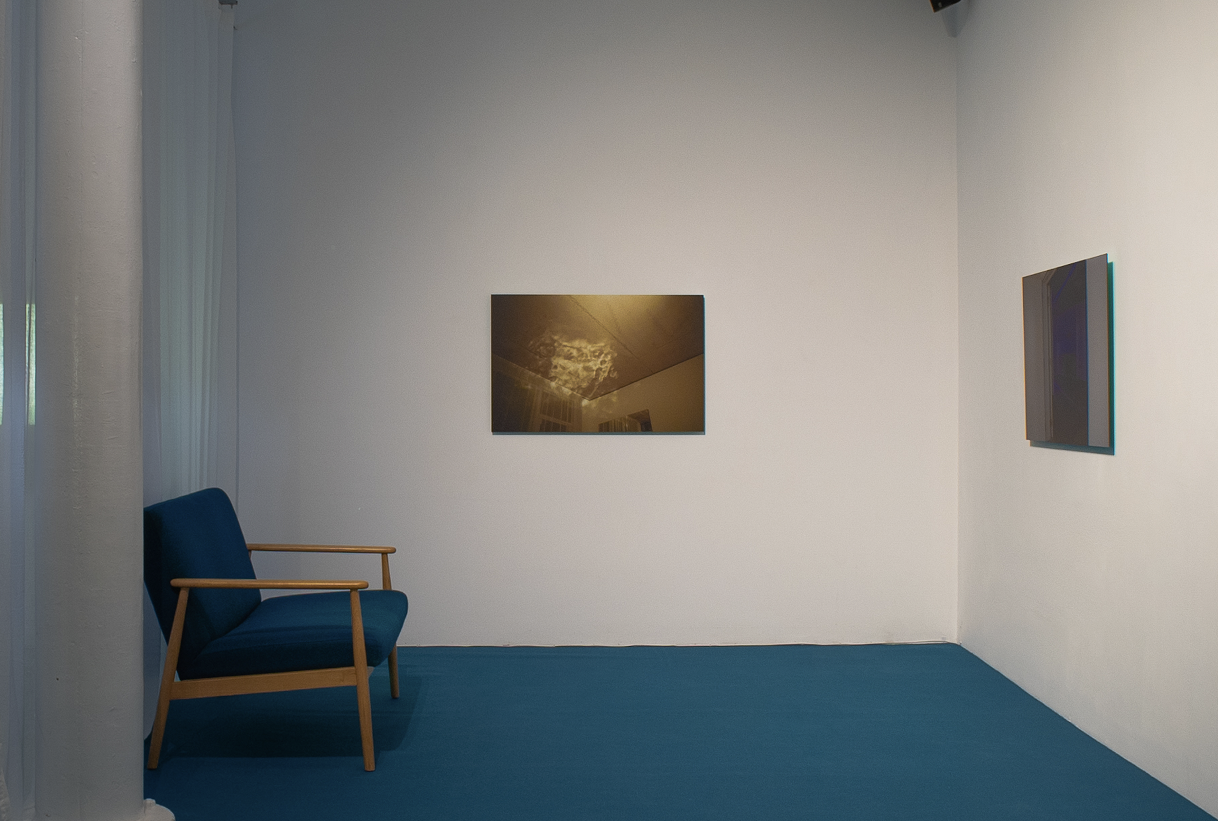 An install shot of an exhibition with two photos on the wall and a seat in the corner, blue carpet on the floor.