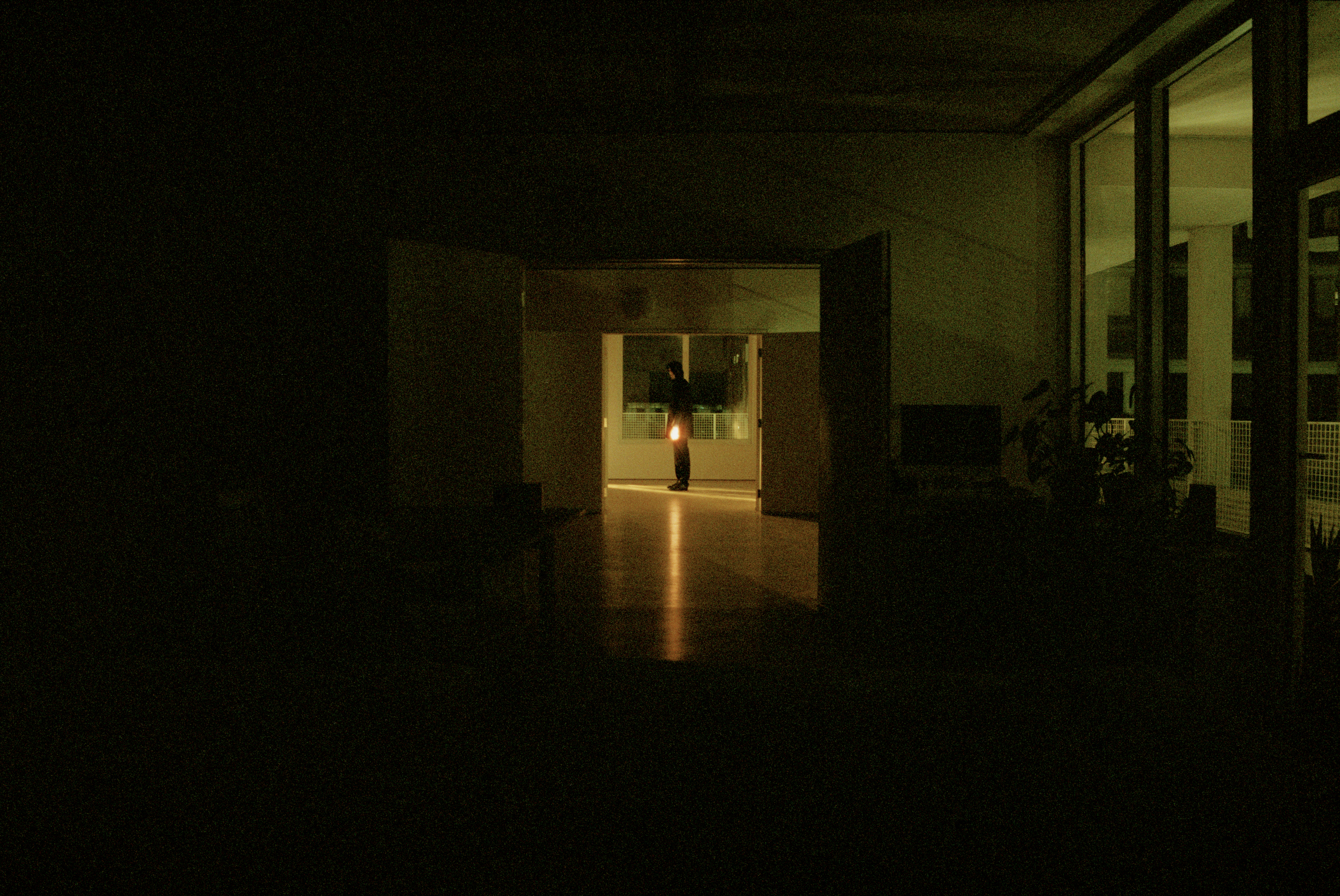 A shadowy view down a long dark room, with a hooded figure standing at the end, with one shining hand.