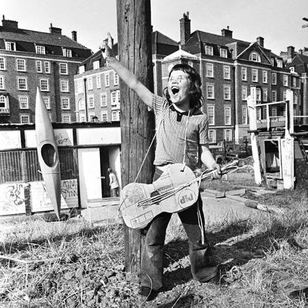 A young girl outdoors on a sunny day, standing next to a telephone pole, smiling and waving, holding a homemade guitar made from cardboard and other materials, with a background of buildings, a playground, and a basketball hoop.