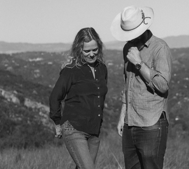 A woman and a man walking outdoors on a hillside, with the woman looking down and the man wearing a wide-brimmed hat and looking at the ground.