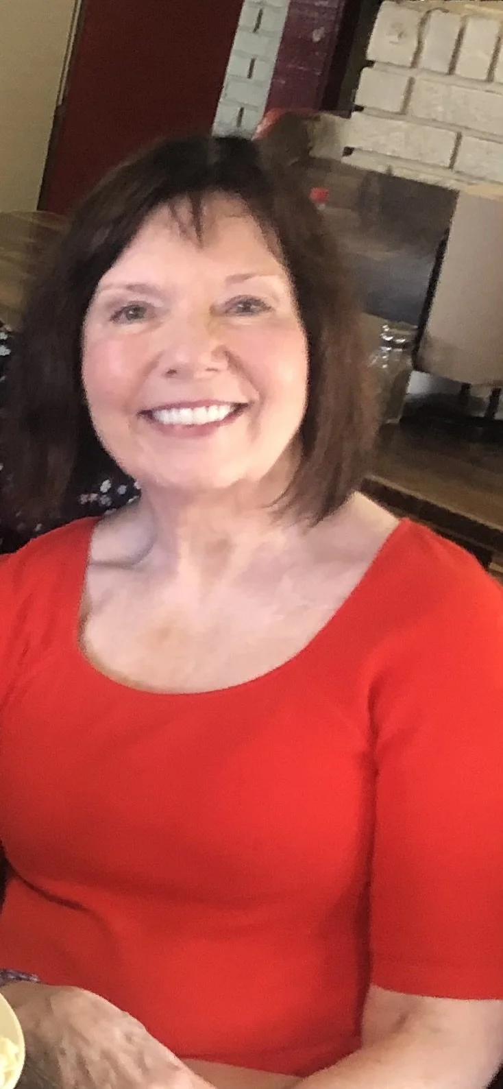 Smiling woman with short brown hair wearing a red top, sitting indoors with a brick wall and furniture in the background.