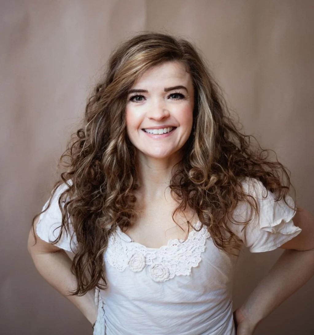 A woman with long, curly brown hair and a bright smile, wearing a white shirt with lace details, stands against a neutral background.