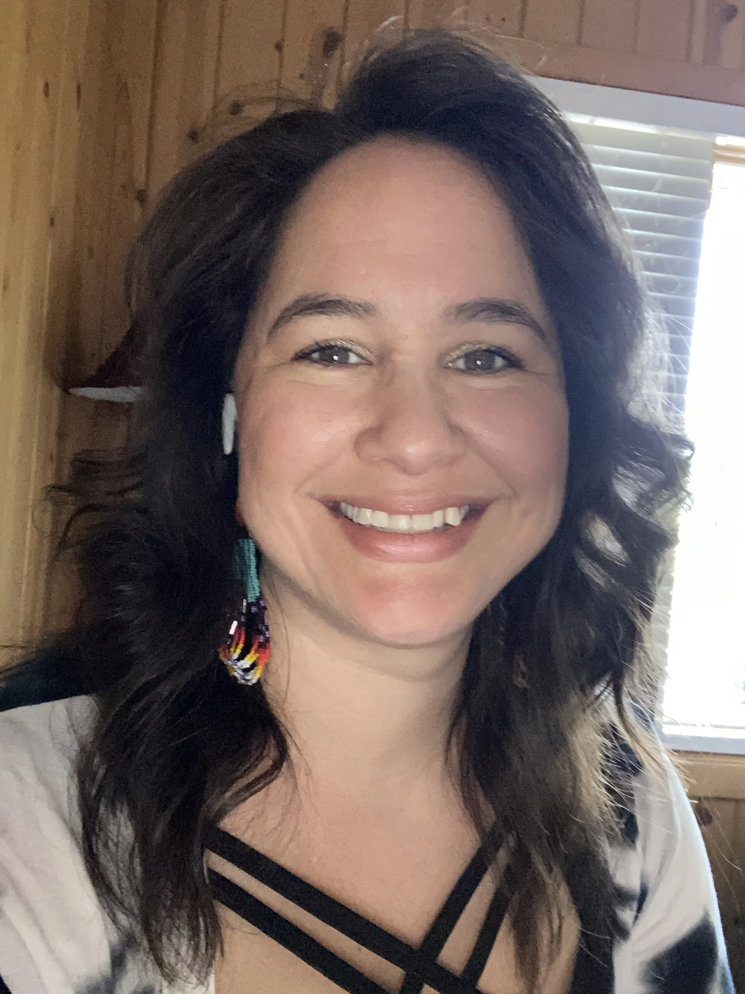 A smiling woman with dark wavy hair, wearing colorful beaded earrings and a white top with black crisscross straps, in front of a wooden wall and a window with blinds.