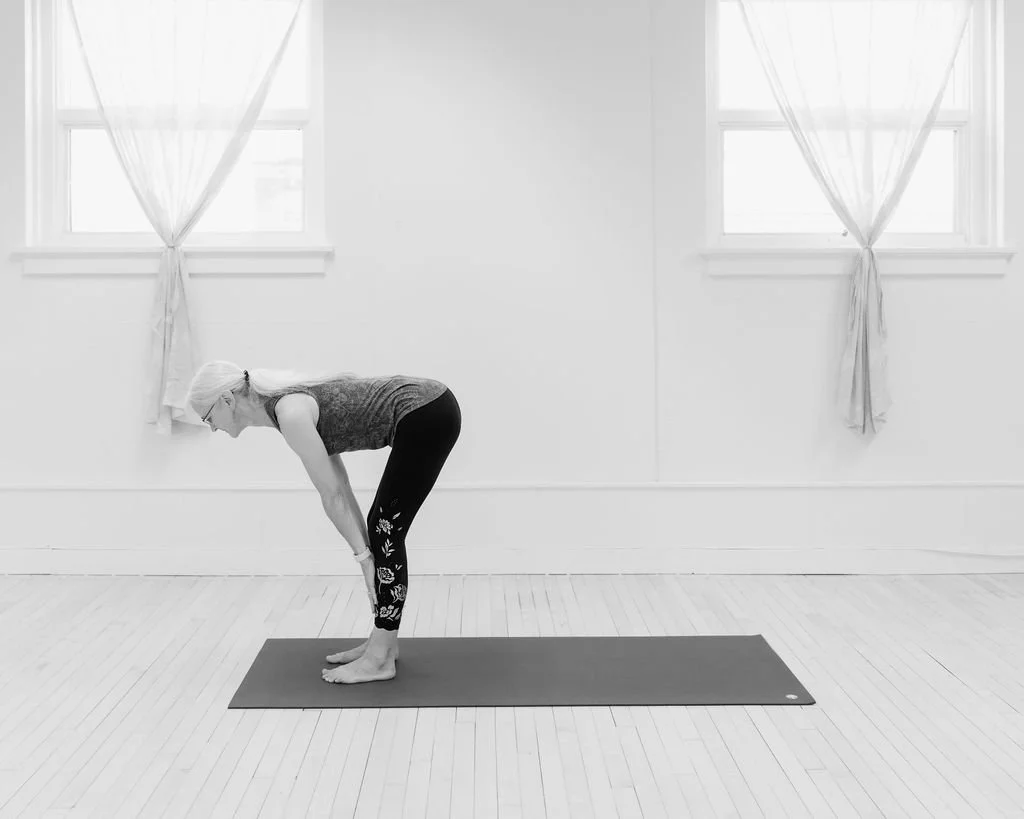 A woman practicing yoga indoors, standing on a yoga mat, bending forward with her hands on her shins.