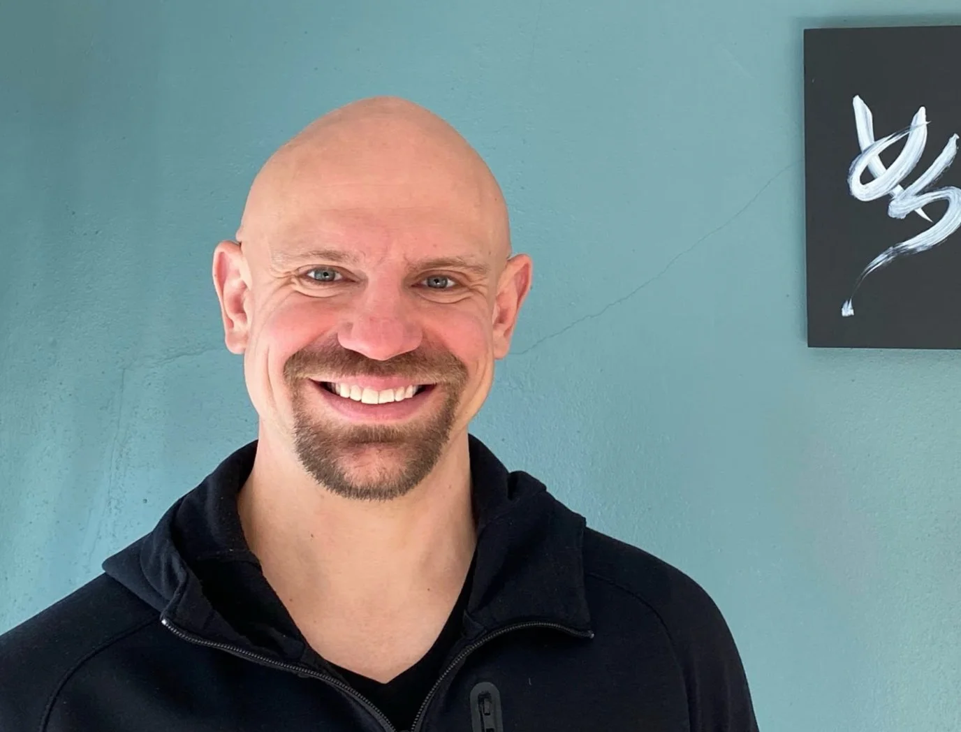 Smiling man with a goatee and bald head, wearing a black zip-up jacket, standing in front of a teal wall with a black and white abstract painting to his right.