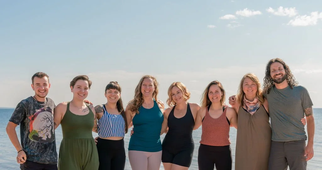 Group of nine friends standing on a beach, smiling with arms around each other under a clear sky.
