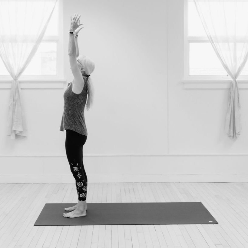 A woman performing yoga in a well-lit room with white walls and large windows, standing on a yoga mat with her arms raised above her head.