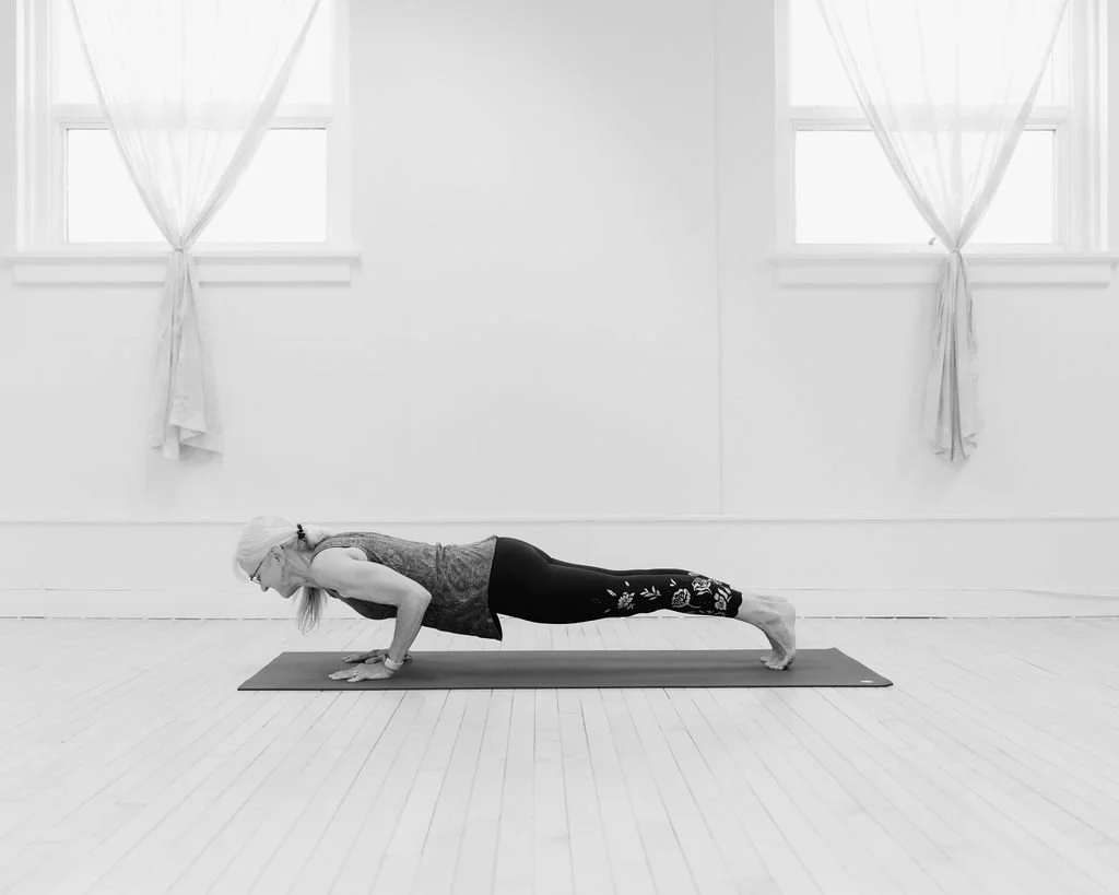 A woman practicing yoga in a plank pose on a mat indoors, with windows and curtains in the background.