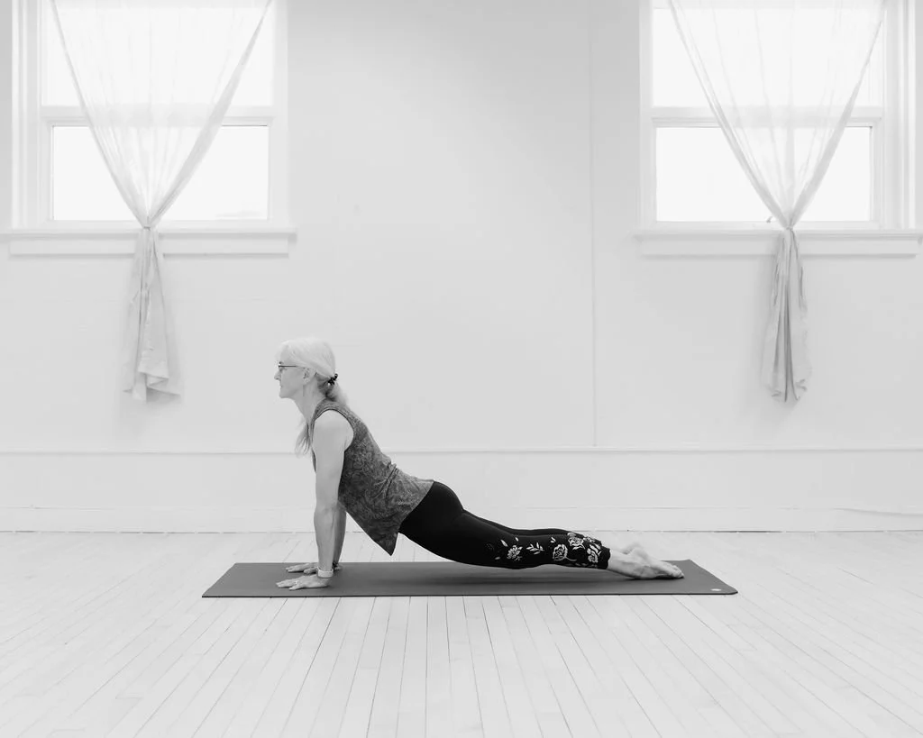 A woman practicing yoga on a mat in a bright room with white walls and large windows, performing a cobra pose.