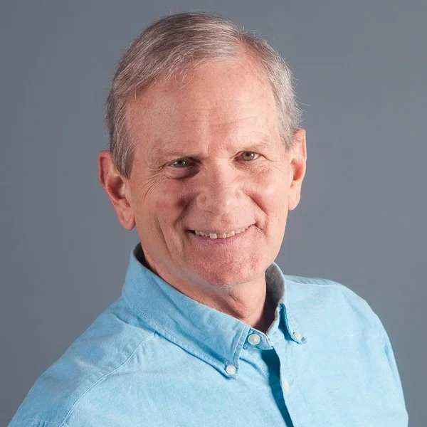 Portrait of a middle-aged man with gray hair smiling, wearing a light blue button-up shirt, against a gray background.