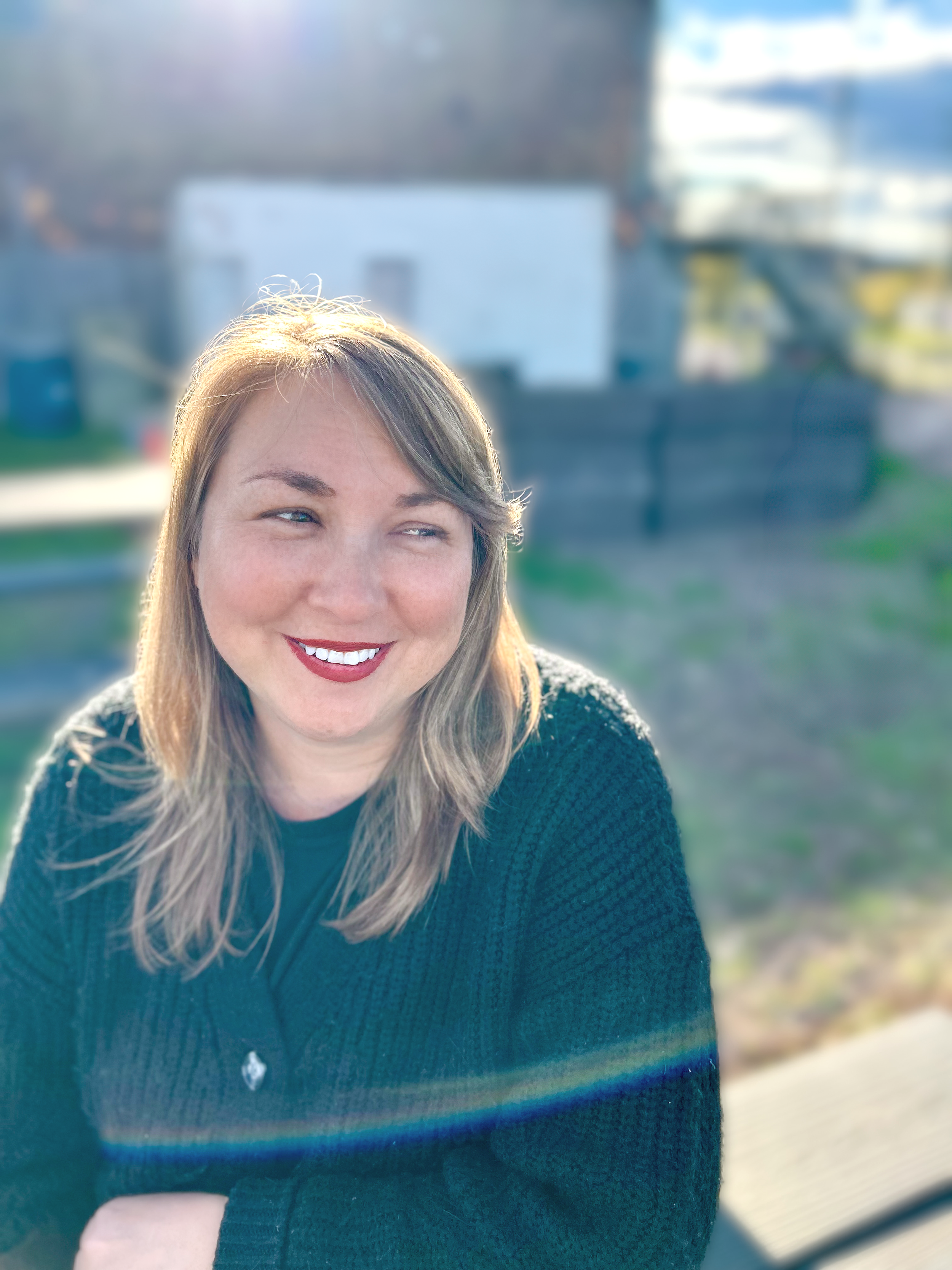 A woman with shoulder-length blonde hair wearing a black sweater, smiling outdoors in sunlight, with blurred background of a backyard and fence.