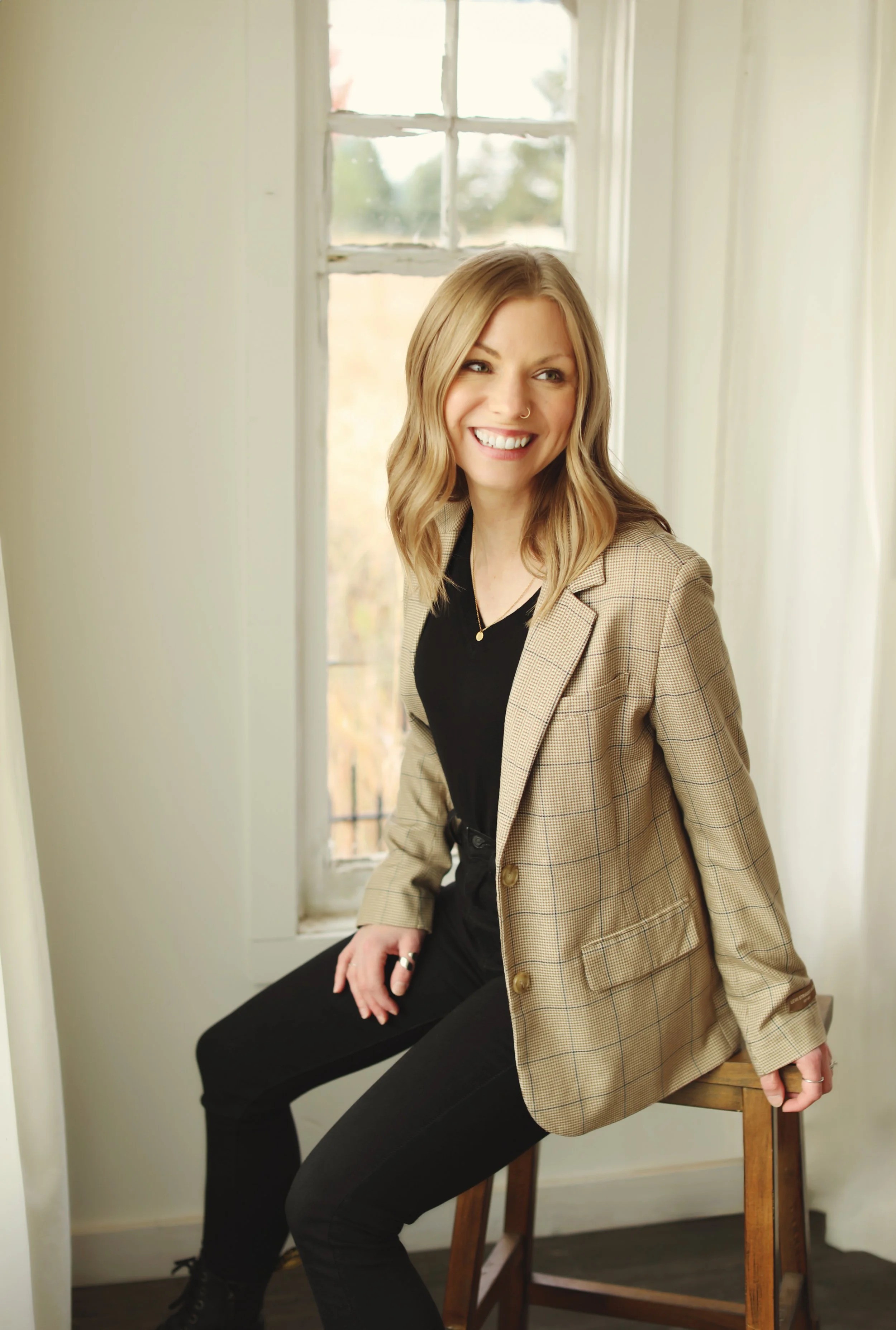 Jessica James, a white woman with green eyes, a gold hoop nose ring, and blonde shoulder length curled hair, sits on a stool smiling and looking away from the camera. She's wearing a brown plaid blazer over a black shirt and pants.