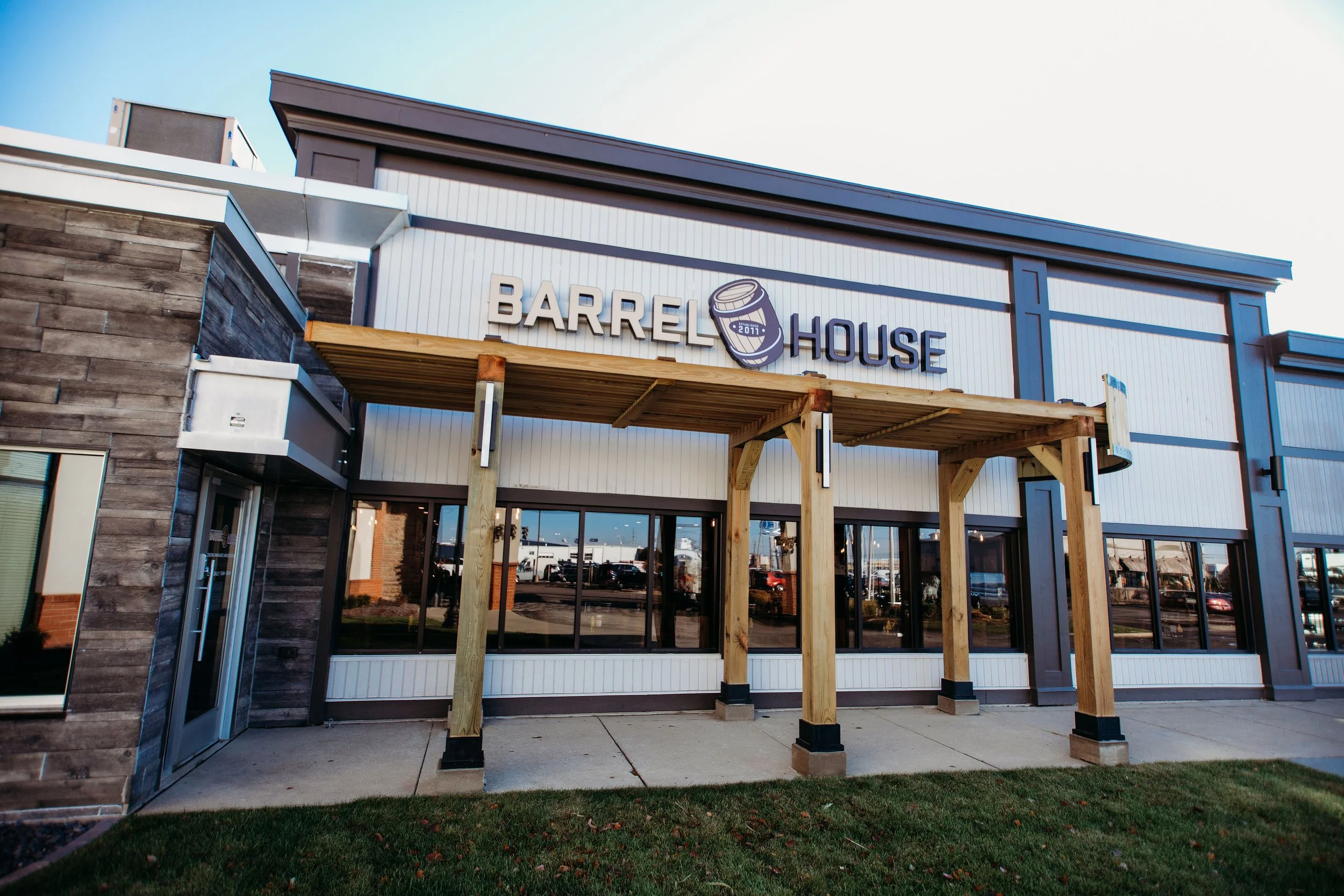 Exterior view of a modern building with a sign reading "Barrel House" and a logo of a barrel, tied to a wooden pergola in front, under a blue sky with some clouds.