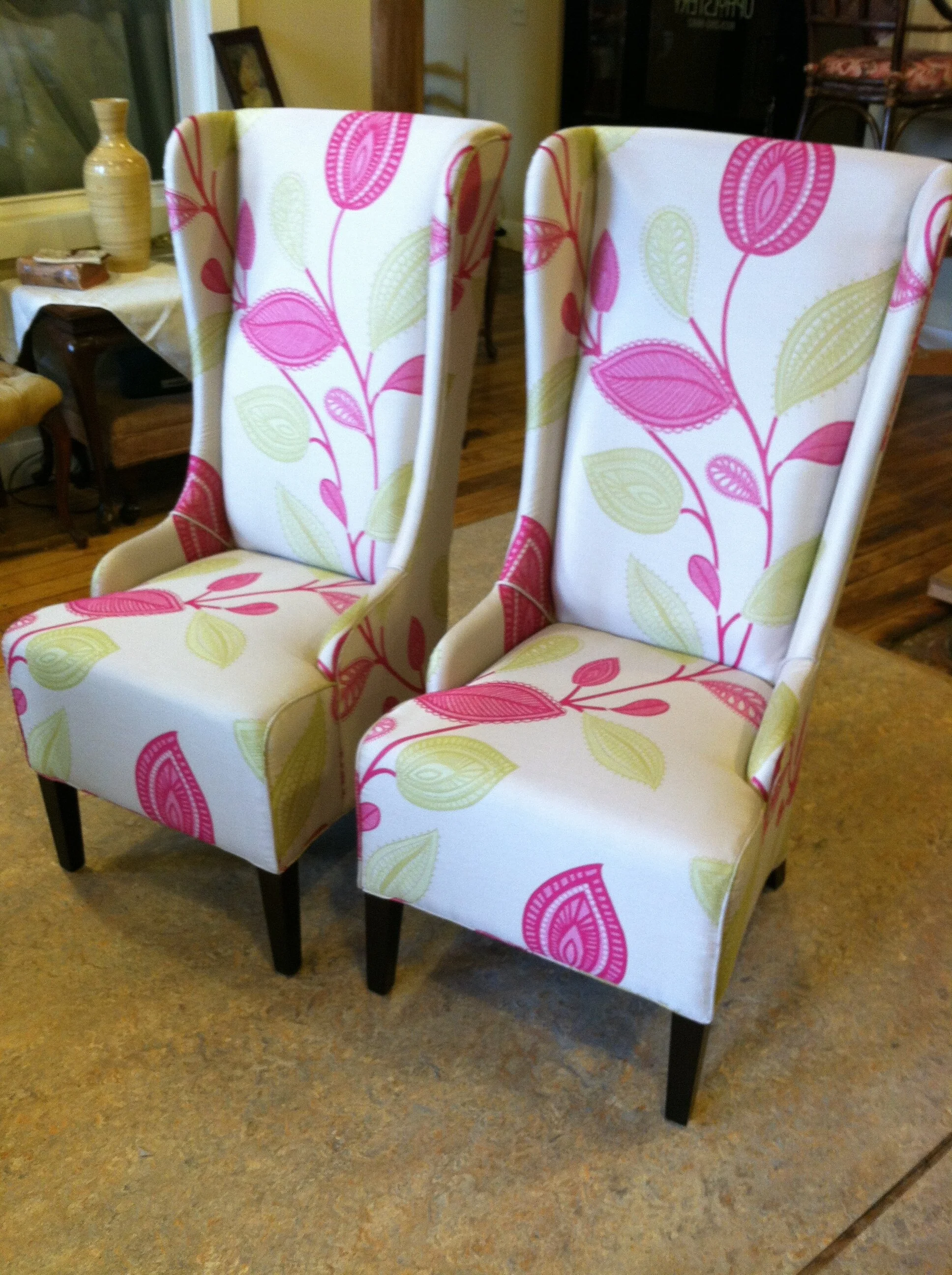 Two upholstered wingback chairs with a white background and pink and green leaf patterns, placed in a room with wooden flooring and various decorative items.