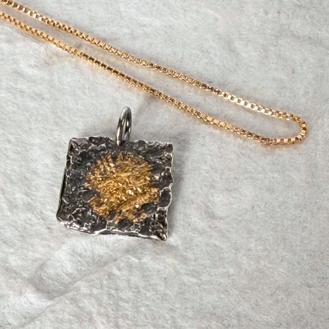 Close-up of the keum-boo gold at the center of the oxidized sterling silver square pendant. The gold sits in the deepest grooves of the radiating texture, catching light against the dark silver.